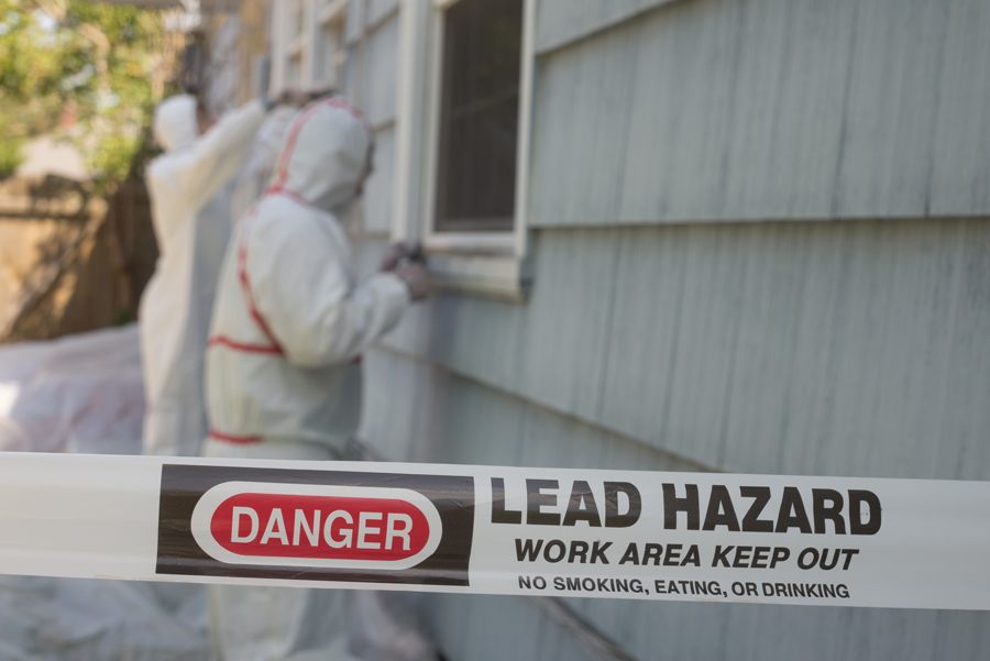 Two people in protective clothing standing next to a house with a sign that says lead hazard, man present.