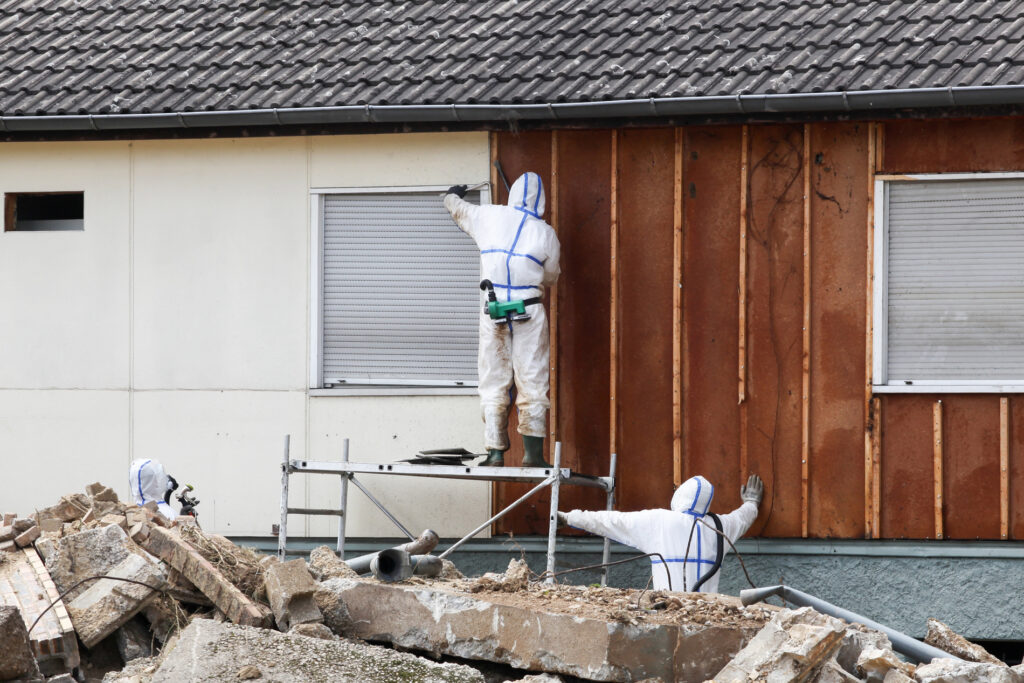 Two men in protective suits working on a house, one man using a drill while the other holds a ladder.