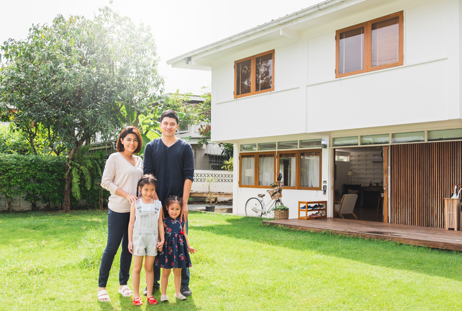 Asian family smiling in front of their new home, a two-story house with a white picket fence.