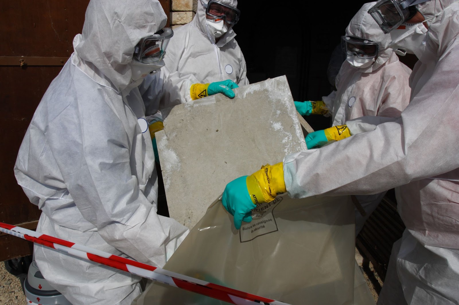 Three men in protective suits and gloves carefully handle a large concrete piece, ensuring safety from asbestos exposure.